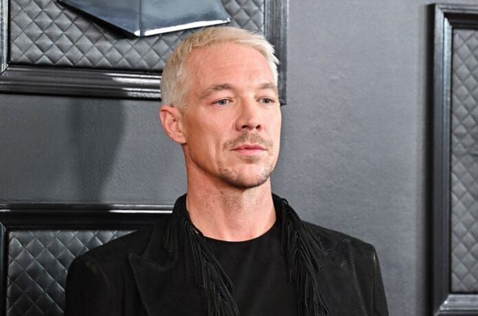 Diplo n a black jacket mourning in front of a black wall.