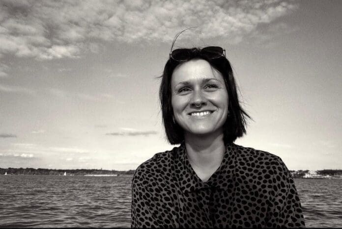 A black and white photo of a woman smiling in front of a body of water.