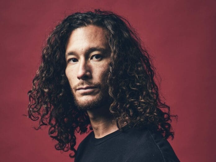 A man with curly hair standing in front of a red background.