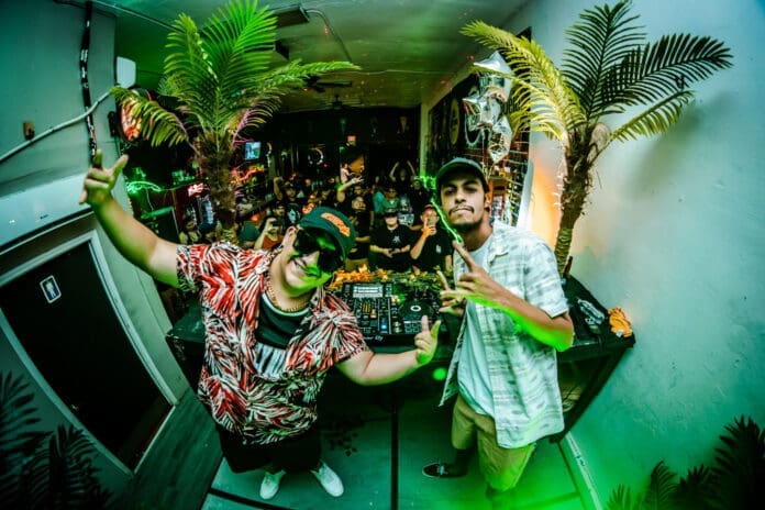 Two men standing in front of a dj booth with palm trees, creating an American Hard Dance Haven.