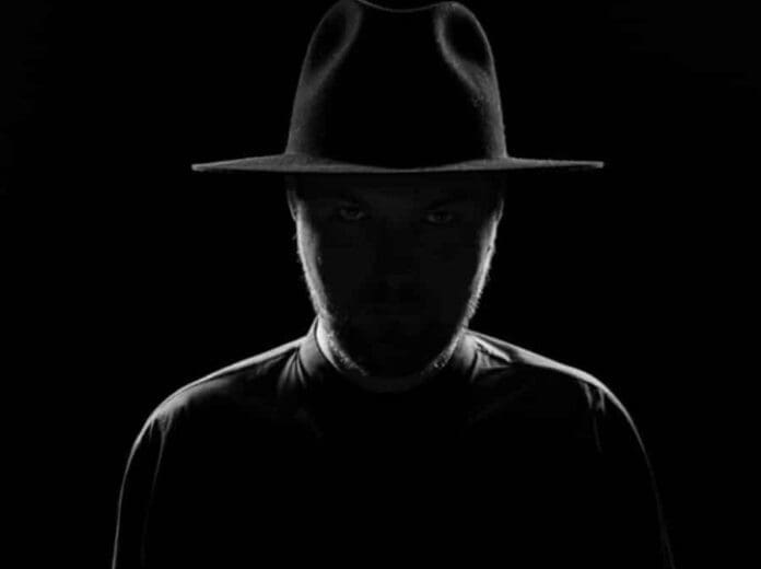 A man in a black hat is standing in front of a black background.