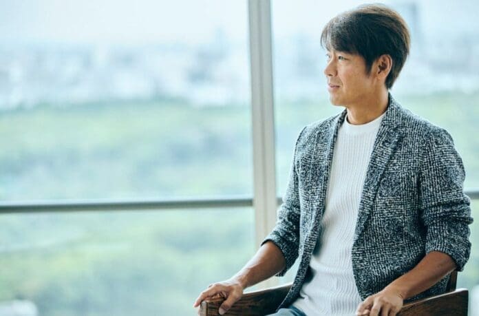 An Asian man, Naoshi Fujikura, in an interview, sitting in a chair in front of a window.