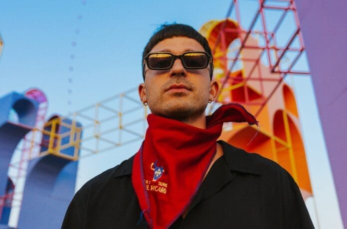 A man in sunglasses and a red bandana walking amid colorful buildings.