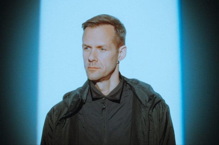 Adam Beyer, wearing a black jacket, stands in front of a vibrant blue light as he unveils his latest EP 'Desert Queen'.