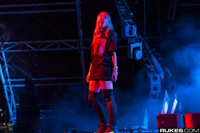 A woman in a black dress standing on a stage, mesmerizing the crowd with her electrifying performance as she brings the sounds of QUIX and Alison Wonderland to life.