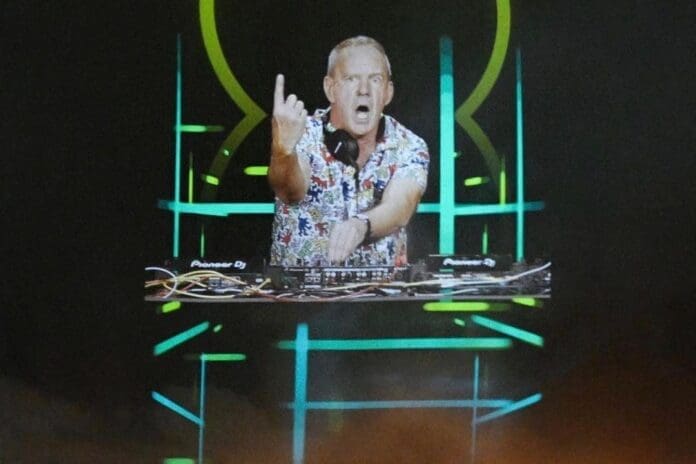 A hologram DJ performing on a stage in front of a large screen.