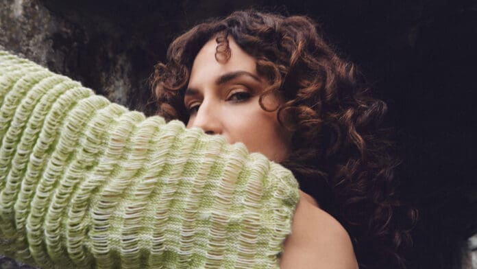 A woman in a green sweater leaning against a La Fleur advertisement on a wall.