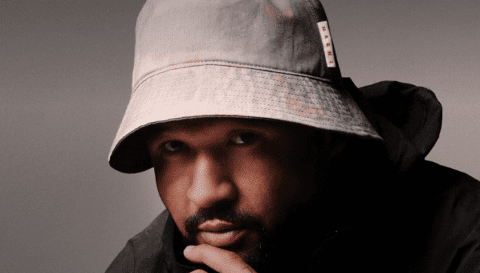 During an interview, a man wearing a bucket hat ponders with his hand on his chin.