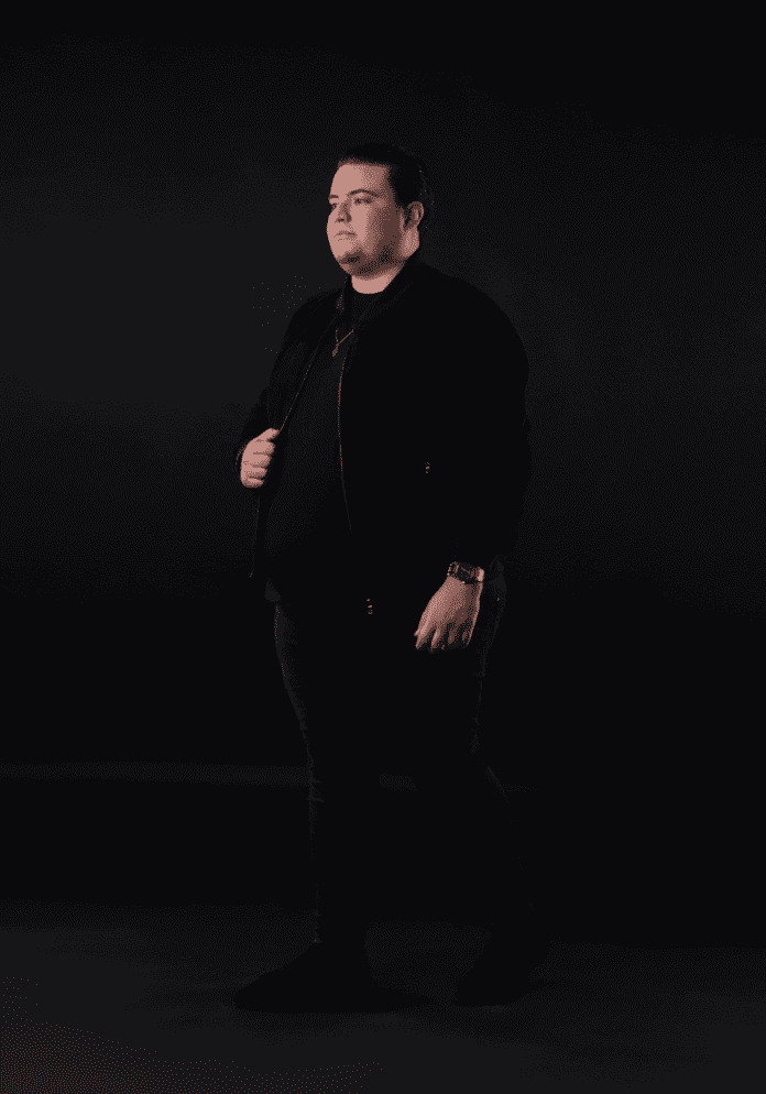 A man in a black jacket is standing in front of a black background while listening to Tony Kay's explosive tech house hit 'Dihya'.