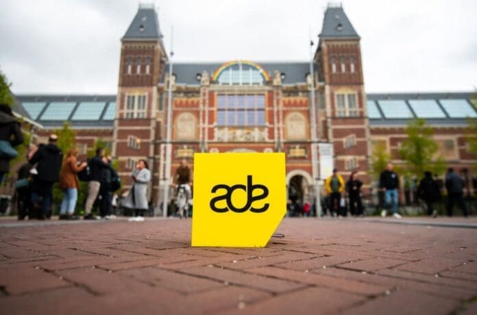 A group of people standing in front of a building with a yellow sign, eagerly discussing their favorite top songs for ADE 2023.