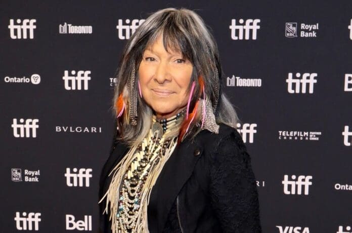 Buffy Sainte-Marie, an Indigenous Heritage icon, gracefully stands on a red carpet, her long hair flowing elegantly as she captures attention and reporters flock to interview her for their upcoming report