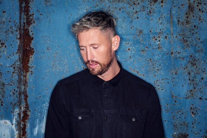 A man in a black shirt standing in front of a blue wall, exuding an anthemic presence.