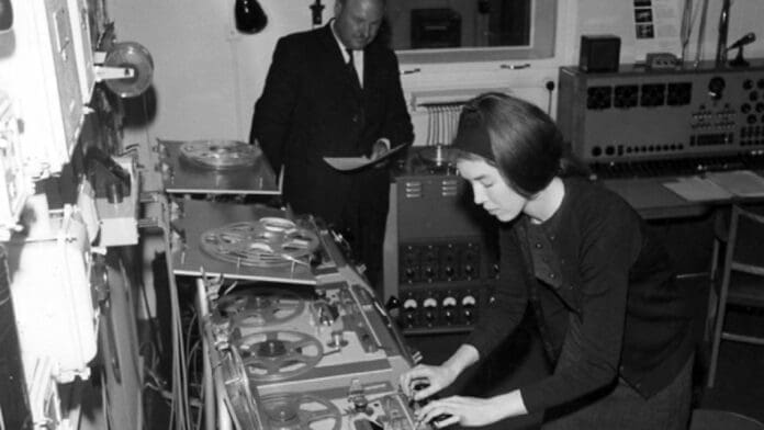 Delia Derbyshire, a woman in a suit and tie, is working on a machine that produces electronic music.