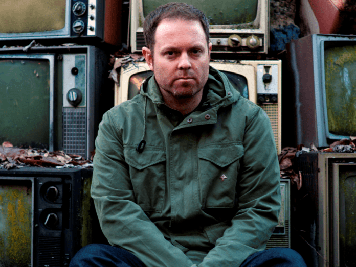 DJ Shadow sitting in front of a pile of TVs while promoting his new album on tour.