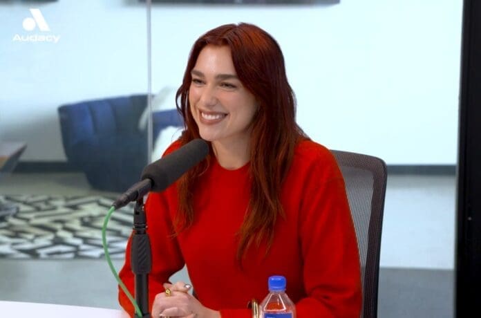 Dua Lipa, wearing a red shirt, is sitting in front of a microphone.