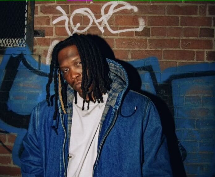 E the Profit, a man with dreadlocks, standing in front of a graffiti wall.