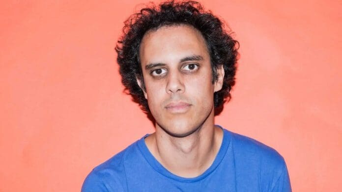 A man with curly hair is standing in front of an orange background at Alexandra Palace.