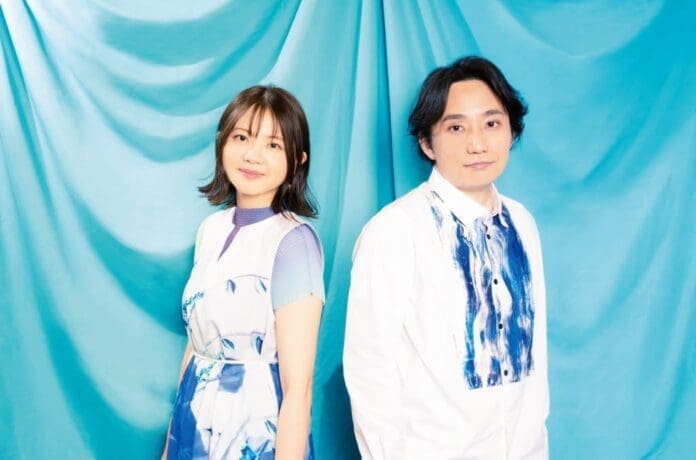 A man and woman, Ureshikute/Tokimeki, posing in front of a blue curtain.