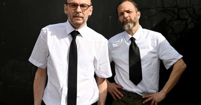 Two men in white shirts and ties standing in front of a black wall promoting their new album 